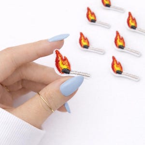 May include: Embroidered patches in the shape of lit matches. The flames are red, orange, and yellow, with a white and gray matchstick. Several patches are scattered on a white background. One patch is held by a hand with light blue nails.