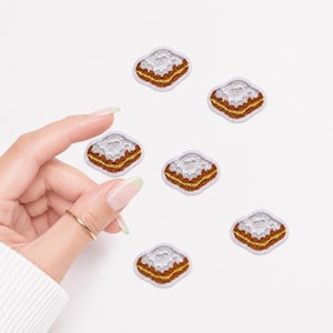 May include: Six small embroidered patches shaped like square pastries with scalloped edges. Each patch features a brown base, a golden-brown middle layer, and a textured silver top, all outlined with a thin white border on a plain white background.