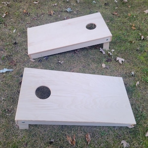 Può includere: Due tavole da cornhole rettangolari realizzate in legno chiaro non rifinito. Ogni tavola presenta una superficie liscia e piatta con un singolo foro circolare posizionato vicino al centro superiore. Le tavole sono inclinate su gambe pieghevoli.