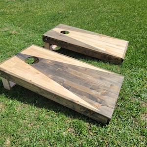 May include: A pair of rectangular wooden cornhole boards featuring a two-tone design with natural light wood and dark stained wood arranged in a triangular pattern. Each board has a circular hole cut into the top surface and sturdy legs.