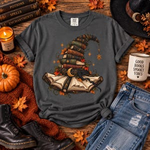 Può includere: Una t-shirt grigio antracite con la grafica di un cappello da strega composto da libri antichi impilati, decorato con foglie autunnali, stelle e una luna crescente. Il design include un libro aperto alla base del cappello in toni caldi.