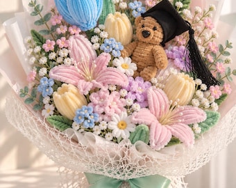 May include: A crochet bouquet featuring a brown teddy bear wearing a black graduation cap, surrounded by blue, pink, and yellow crocheted flowers, green leaves, and white filler flowers, wrapped in white paper with a light green ribbon.