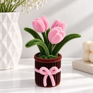 May include: A decorative potted plant featuring three pink tulips crafted from fuzzy pipe cleaners. The tulips have green stems and leaves, and they are set in a dark brown, ribbed cylindrical pot adorned with a light pink pipe cleaner bow.