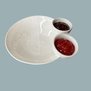 May include: White ceramic serving dish with two attached condiment cups, one filled with a dark sauce and the other with red sauce. The dish is on a light blue background.