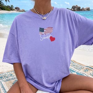 Könnte beinhalten: Ein hellviolettes Kurzarm-T-Shirt mit einer Grafik einer verwaschenen amerikanischen Flagge über dem weißen, kursiven Schriftzug "Sweet" und einem kleinen roten Herz. Das T-Shirt hat einen Rundhalsausschnitt und eine lockere, entspannte Passform.