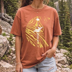 Può includere: Una t-shirt a maniche corte color ruggine con una grande grafica gialla a forma di mezzaluna riempita di motivi vorticosi e un pianeta centrale con anelli. La t-shirt è abbinata a pantaloncini di jeans azzurri con uno strappo sulla coscia.