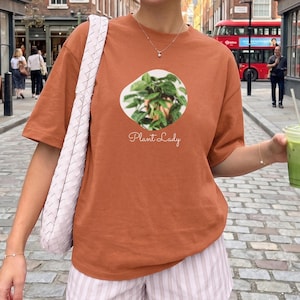 以下が含まれることがあります： 錆色の半袖Tシャツで、緑色の鉢植えの円形グラフィックと、その下に白い筆記体でPlant Ladyという文字がプリントされています。このTシャツは、薄いピンクと白の縦縞模様のショートパンツと組み合わされています。