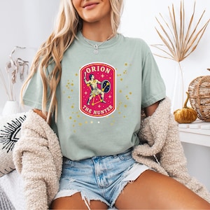 May include: A sage green short-sleeve t-shirt featuring a central graphic of the Orion constellation depicted as a hunter with a club and shield. The design is enclosed in a pink oval border with the text ORION THE HUNTER and yellow stars.