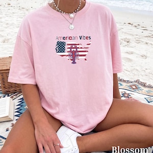Puede incluir: Una camiseta de manga corta de color rosa claro con un estampado gráfico del mapa de los Estados Unidos relleno con el patrón de la bandera estadounidense, una ilustración de una langosta roja y el texto American Vibes en letras negras y rojas.