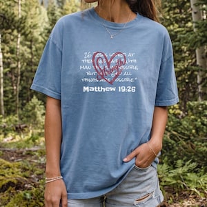 May include: A blue short-sleeved t-shirt featuring a red heart graphic with the word Strength inside. The text reads: 26 Jesus looked at them and said, "With man this is impossible, but with God all things are possible." Matthew 19:26.