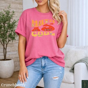 Op de afbeelding: Een roze T-shirt met korte mouwen en de gele en oranje tekst "MATH IS COOL" op de borst. De afbeelding bevat wiskundige symbolen en formules zoals "e=mc2" en "a2+b2=c2" die in het ontwerp zijn verwerkt. Het T-shirt heeft een ronde hals.