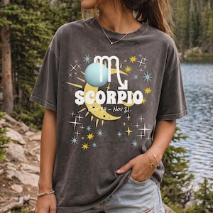 May include: A charcoal grey short-sleeve t-shirt featuring a graphic print of the Scorpio zodiac symbol, a crescent moon, a light blue sphere, and yellow and white stars. The text reads SCORPIO and Oct 24 - Nov 21 in white and yellow lettering.