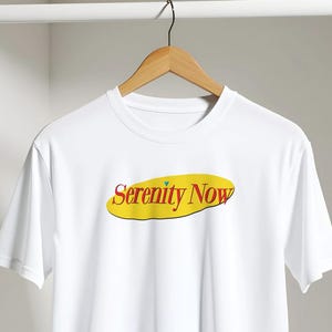 May include: White t-shirt with the text "Serenity Now" in red and yellow. The text is inside a yellow oval. The t-shirt is hanging on a wooden hanger.