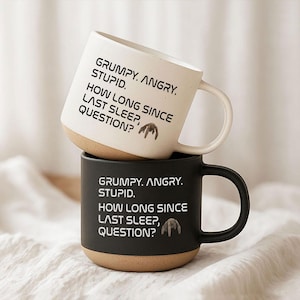 May include: Two ceramic mugs, one white and one black, stacked on top of each other. Each mug has a brown base and a curved handle. The white mug has the text "GRUMPY. ANGRY. STUPID. HOW LONG SINCE LAST SLEEP, QUESTION?" printed in black. The black mug has the same text in white.