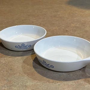 May include: Two white, oval-shaped serving dishes with handles. Each dish has a blue floral design on the side. The dishes are on a brown countertop.