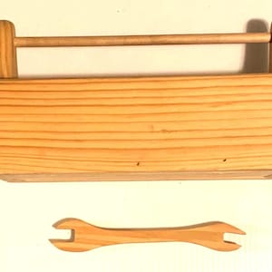 May include: A rectangular light-colored wooden toolbox featuring a horizontal handle spanning the top between two vertical supports. Below the toolbox lies a small, flat, double-ended wooden wrench with an open-ended design on both sides.