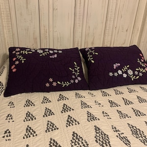 May include: Two rectangular deep purple quilted pillow shams featuring embroidered floral and butterfly motifs in shades of pink, white, and light green, positioned on a white bedspread with a repeating gray triangular geometric pattern.