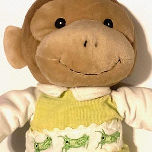 May include: A plush monkey toy with brown fur, black eyes, and a stitched smile. The monkey wears a white collared shirt under a light green vest featuring a white smocked panel decorated with embroidered green grasshopper illustrations.