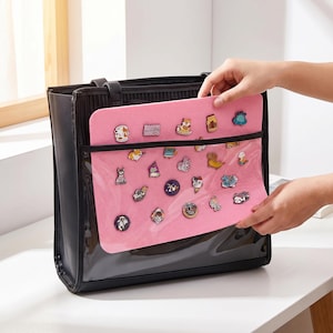 Puede incluir: Bolso tote negro con un bolsillo frontal transparente que contiene una hoja de fieltro rosa cubierta de alfileres decorativos. Los alfileres presentan varios diseños de animales y objetos. Una mano sostiene la hoja rosa.