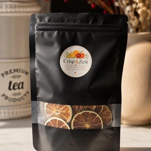 May include: Black bag of dried citrus fruit slices with a clear window. The bag has a circular label with the text "Crisp & Zest" and illustrations of citrus fruits. The bag is on a white surface.