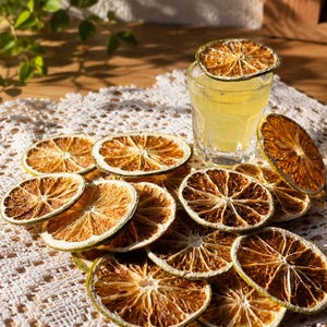 Peut inclure: Un verre de liquide jaune avec une tranche d'agrume séchée sur le dessus, entouré de plusieurs tranches d'agrumes séchées. L'arrangement repose sur un napperon en dentelle blanche sur une surface en bois. Des feuilles vertes sont visibles en arrière-plan.