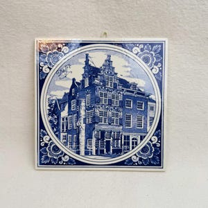 May include: Square ceramic tile with a blue and white Delft design. The tile features a detailed illustration of a building within a circular frame, surrounded by floral patterns. The word "Delft" is written in script.