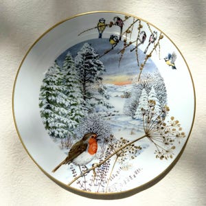 May include: A white decorative plate with a gold rim featuring a winter scene with snow-covered trees, a red-breasted robin, and other birds perched on branches. The plate has a detailed, painted design.
