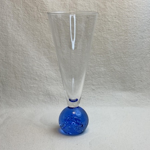 May include: A clear glass champagne flute with a vibrant blue, spherical base. The glass has a long, slender stem and a wide, conical bowl. The blue base is filled with small air bubbles, adding a decorative touch to the elegant glassware.