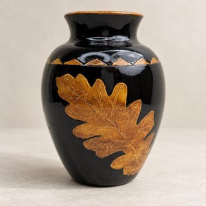 May include: A glossy black ceramic vase featuring a prominent textured brown oak leaf relief on the front. The vase has a rounded body, a narrow neck, and a wide rim with a brown glazed edge. A decorative band of brown triangles encircles the neck.