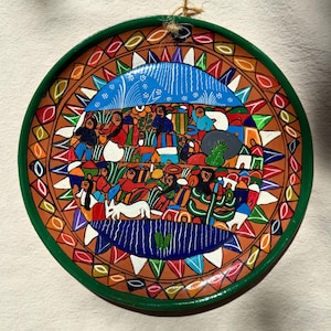 May include: A hand-painted decorative plate with a colourful design. The plate has a green rim and a central scene with figures, animals, and geometric patterns in orange, blue, red, and white. A jute string is attached for hanging.