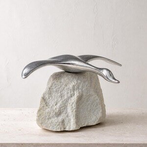 May include: A polished silver-toned metal sculpture of a bird in flight, featuring smooth, elongated wings and a curved neck, mounted securely atop a rough, light grey natural stone base with a jagged, uneven texture and irregular shape.
