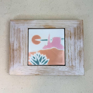 May include: A framed ceramic tile with a desert landscape design. The artwork shows a pink rock formation, a setting sun, and a blue-green plant against a peach and white backdrop. The frame is made of light-coloured, distressed wood.