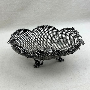 May include: A silver-toned metal bowl featuring a delicate lattice-work design. The rim and the three feet are decorated with detailed relief carvings of grape clusters and vine leaves, creating a textured appearance throughout the piece.