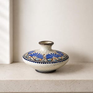 May include: A low-profile, wide-bodied ceramic vase with a speckled cream base. The exterior features hand-painted blue floral motifs, brown scrolling vines, and a dark blue dotted border around the midsection, finished with a dark brown rim.