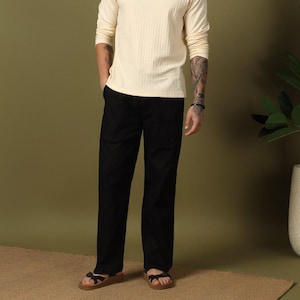 Op de afbeelding: Een persoon draagt een crèmekleurig shirt met lange mouwen en een kraag en een zwarte broek. Het shirt heeft een geribbelde textuur. De persoon draagt sandalen. De achtergrond is effen olijfgroen.