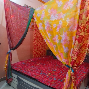 Puede incluir: Una cama con un edredón rojo estampado y un dosel de tela colorida. El dosel presenta tela amarilla con estampados florales y tela roja con estampados florales. El marco de la cama es gris.