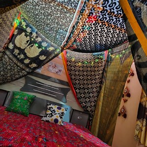 Puede incluir: Un interior de dormitorio con una colcha roja con estampado floral, cojines verdes y estampados, y un dosel hecho de telas estampadas. El dosel está drapeado sobre la cama, creando un espacio único. Luces de hadas están colgadas alrededor del dosel.