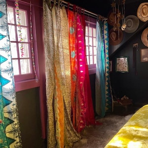 Könnte beinhalten: Eine Reihe von bunten Stoffvorhängen vor Fenstern mit Mustern in Türkis, Gold, Orange und Pink. Der Raum verfügt über eine schwarze Wand, die mit verschiedenen Hüten, einem halbmondförmigen Regal und Wandkunst dekoriert ist.