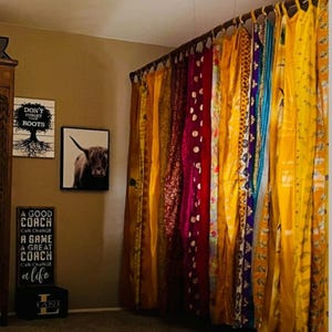 Puede incluir: Una serie de paneles de tela coloridos colgados de una barra, con varios patrones y tonos que incluyen amarillo, rojo y morado. En la pared, tres letreros enmarcados muestran el texto: Don't Forget Your Roots, una vaca highland y A Good Coach.