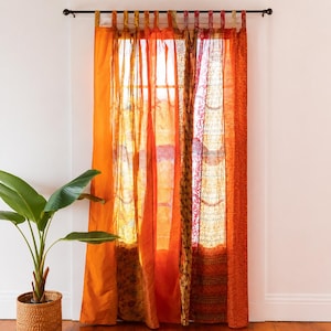 May include: A set of tab-top curtains featuring a patchwork of sheer and opaque fabrics in various shades of orange, gold, and red. The panels display a mix of solid colours and intricate patterns, hanging from a dark metal rod over a window.