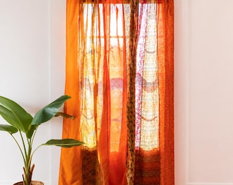 May include: A set of tab-top curtains featuring a patchwork of sheer and opaque fabrics in various shades of orange, gold, and red. The panels display a mix of solid colors and intricate patterns, hanging from a dark metal rod over a window.
