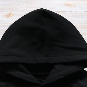 May include: A close-up shot of a black hoodie hood. The hood is made of a soft, dark fabric and is positioned against a light-colored wooden background. The hood is folded over, creating a layered effect.