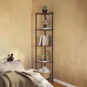 May include: A dark brown corner shelf unit with four shelves. The shelves hold books, plants, decorative items, and a woven basket with rolled towels. The unit has a metal frame and is placed in a corner against a light brown wall.