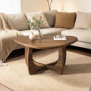 May include: A wooden, triangular coffee table with a dark brown finish. The table has a unique, curved base and is placed on a light-colored rug. A vase with greenery and a small decorative vase are on the table.