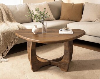 May include: A wooden, triangular coffee table with a dark brown finish. The table has a unique, curved base and is placed on a light-colored rug. A vase with greenery and a small decorative vase are on the table.