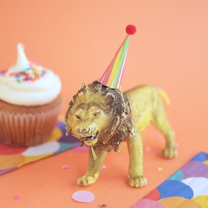 May include: A golden lion figurine wearing a party hat, next to a cupcake with white frosting and sprinkles. The scene is set on an orange surface with confetti and colorful paper. A festive birthday theme.