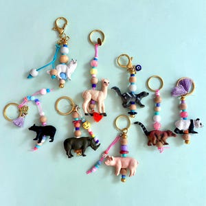 May include: A collection of eight keychains featuring miniature animal figurines including a bear, rhino, pig, panda, llama, and skunk. Each keychain is attached to a gold-tone metal ring and decorated with colorful plastic, wooden, and metallic beads.