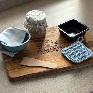 May include: A wooden chopping board with a jar covered in floral fabric, a small blue bowl with a cloth, a black square baking tin, a wooden spatula, and a blue crocheted pot holder. The board has the text "THE MOMMA BEHR SHOP" and a bear logo.