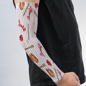 May include: An arm sleeve featuring a white background with thin vertical black pinstripes. The pattern includes illustrations of brown baseball gloves, wooden bats, white baseballs, and red baseball caps, with the word Baseball in red script.