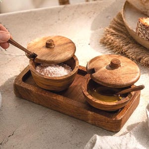 May include: A wooden salt and pepper set with lids and small spoons, presented on a wooden tray. One bowl contains pink salt. The set is next to a plate of bread, suggesting a dining or kitchen theme.